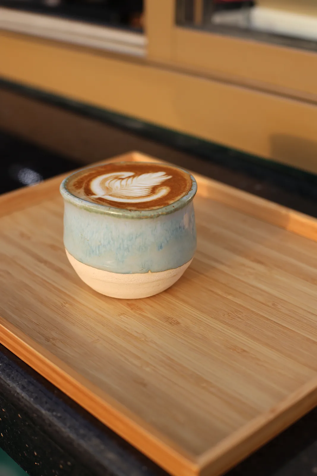 Handmade ceramic cup with a pale blue-grey glaze and a bare clay foot, holding a latte with leaf art, on a bamboo serving tray.