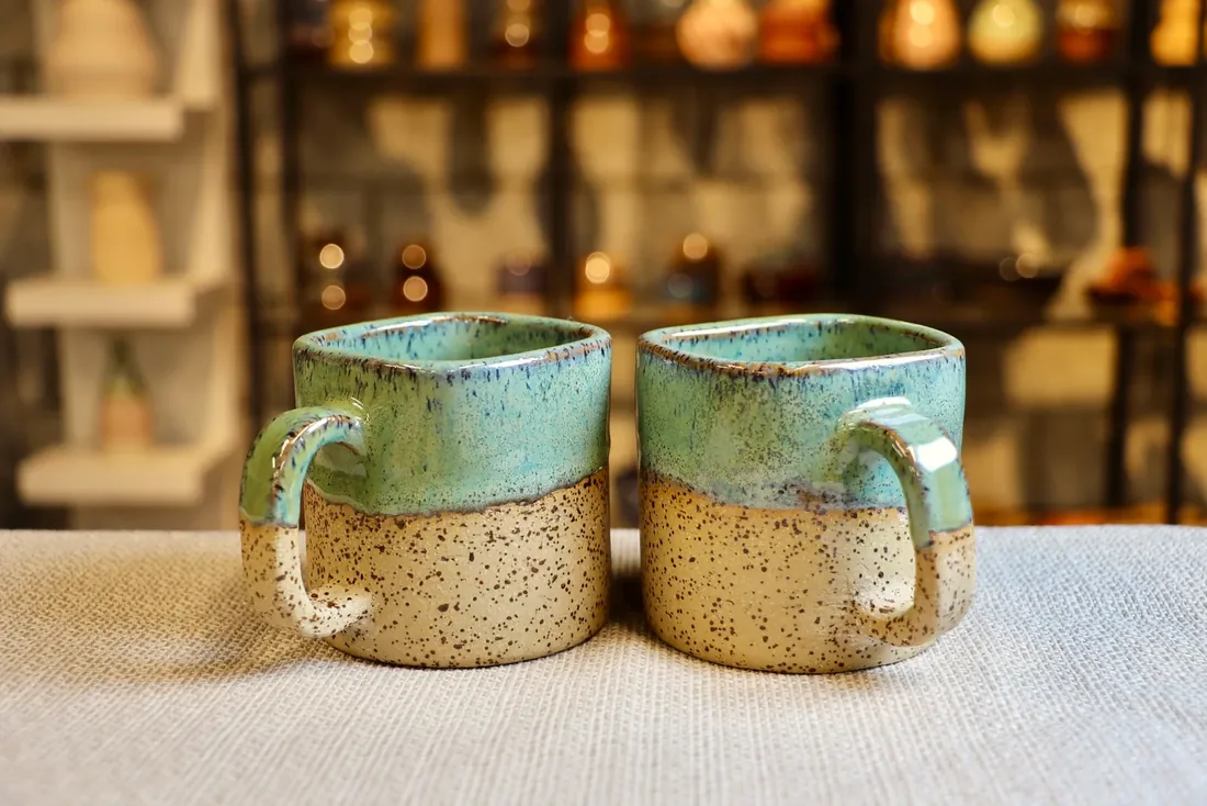 A pair of speckled ceramic cups with handles and a sea-green glaze, side by side on a linen cloth.
