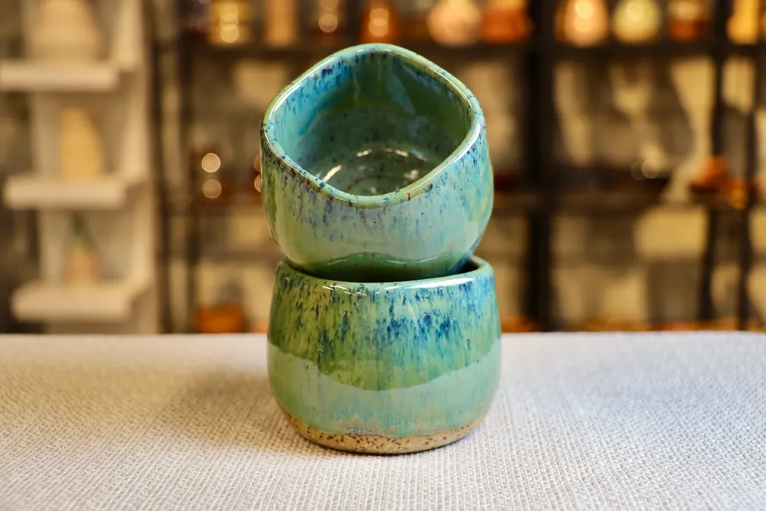 Two speckled ceramic cups with a green and blue glaze, one stacked on the other, on a linen cloth.