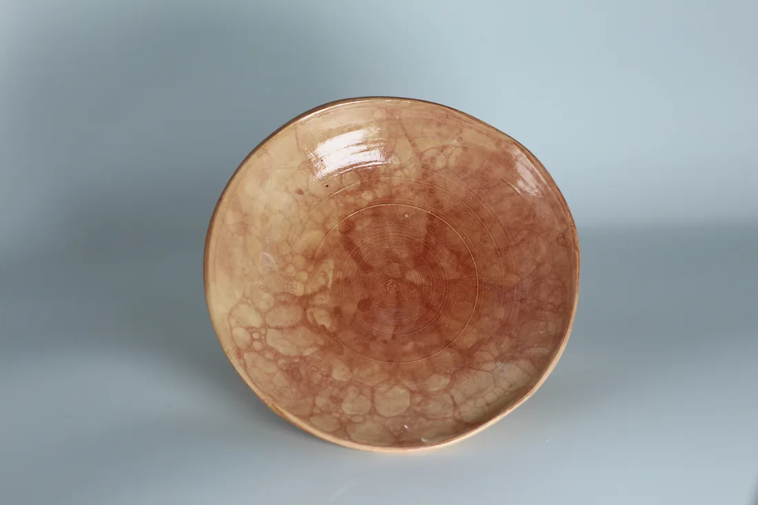 A shallow dish with a pink-amber bubble glaze, seen from above, on a pale grey-blue backdrop.