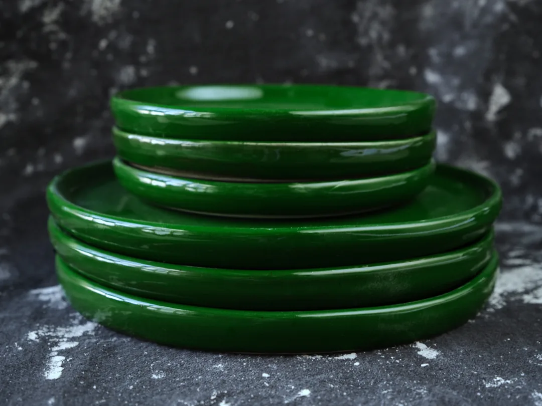 A stack of glossy deep-green plates, three smaller on top of three larger, seen from the side on a dark cloth.