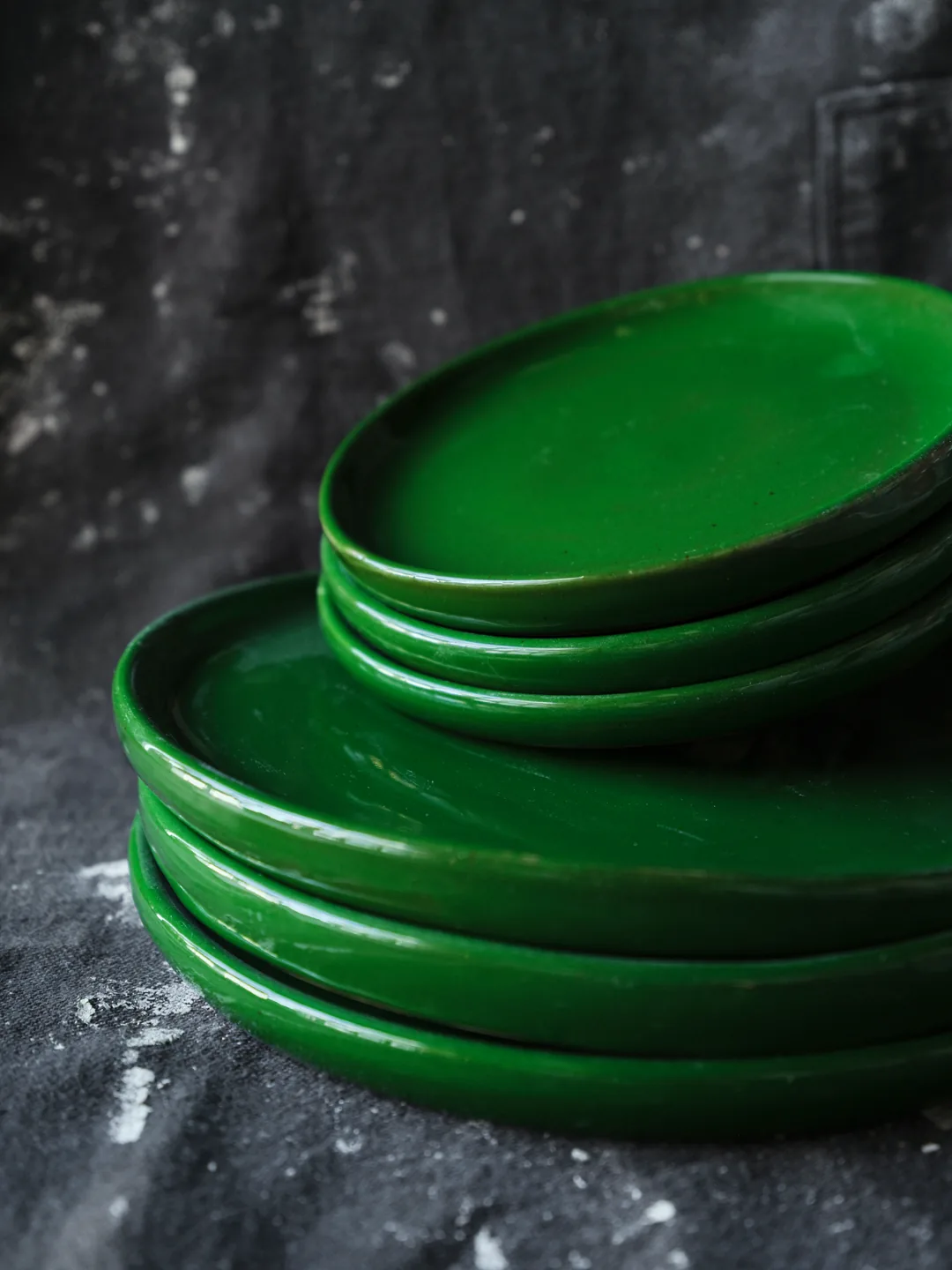 A stack of glossy deep-green handmade ceramic plates in two sizes on a dark cloth.