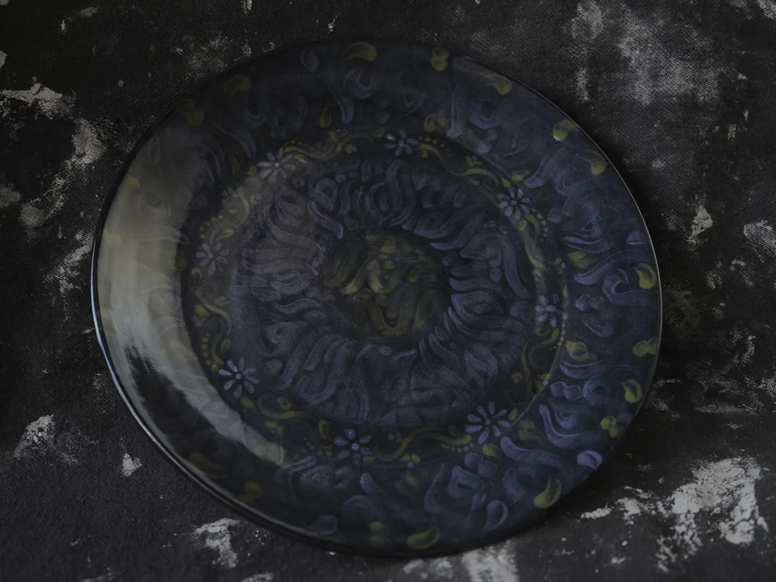 A round plate in dark blue with an embossed pattern of flowers and swirls, on a dark cloth.