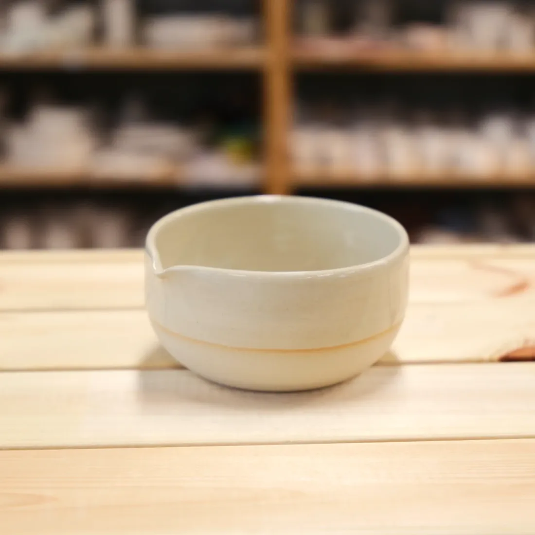 A plain cream handmade spouted bowl on a light wood table.