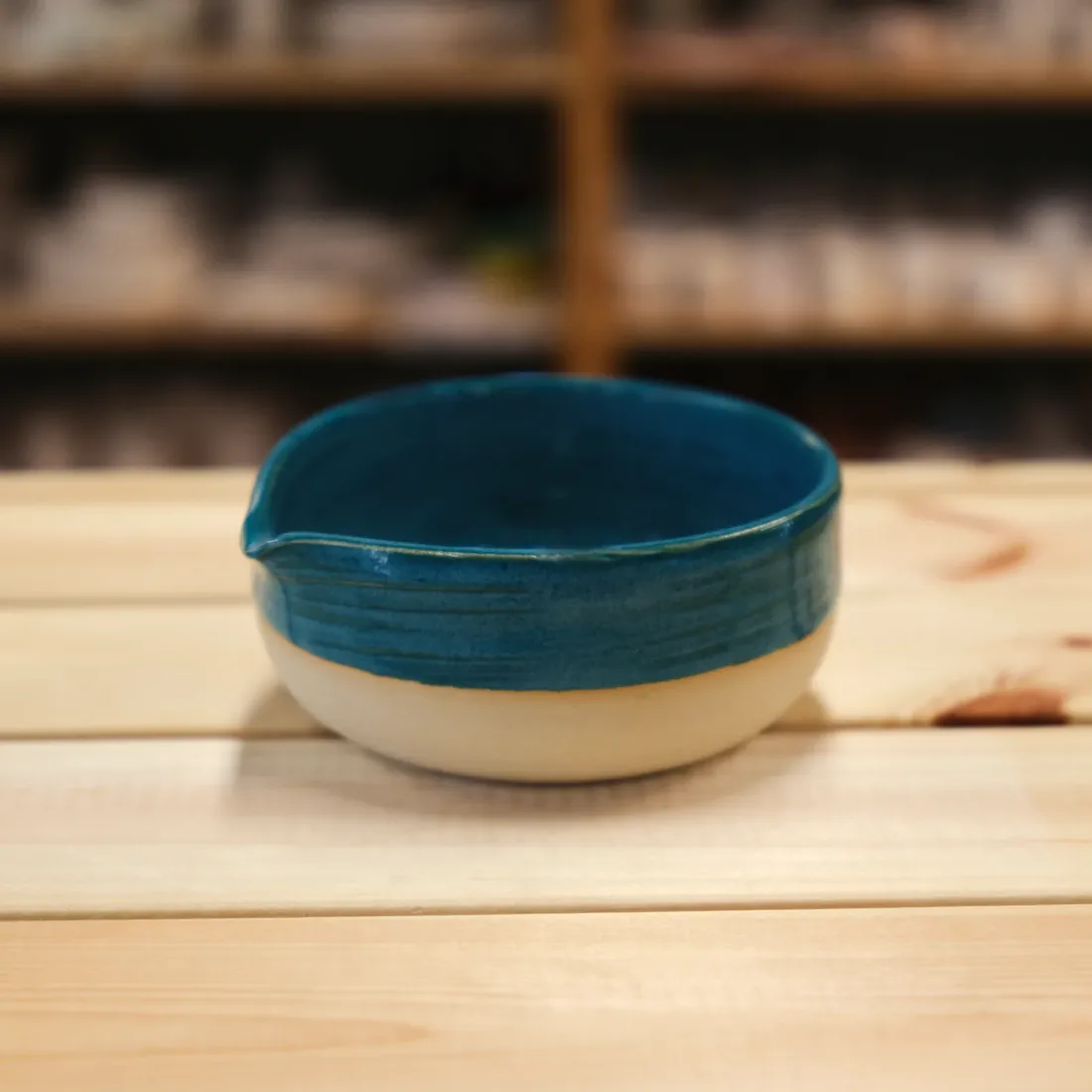 A handmade spouted bowl with a deep teal glaze over a cream base, on a light wood table.