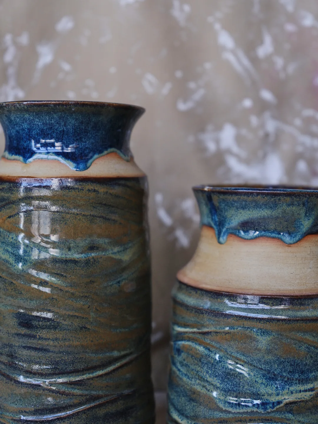 Close-up of two vases with a wavy textured surface and a layered blue-green glaze, with a band of bare clay below a blue-glazed rim.