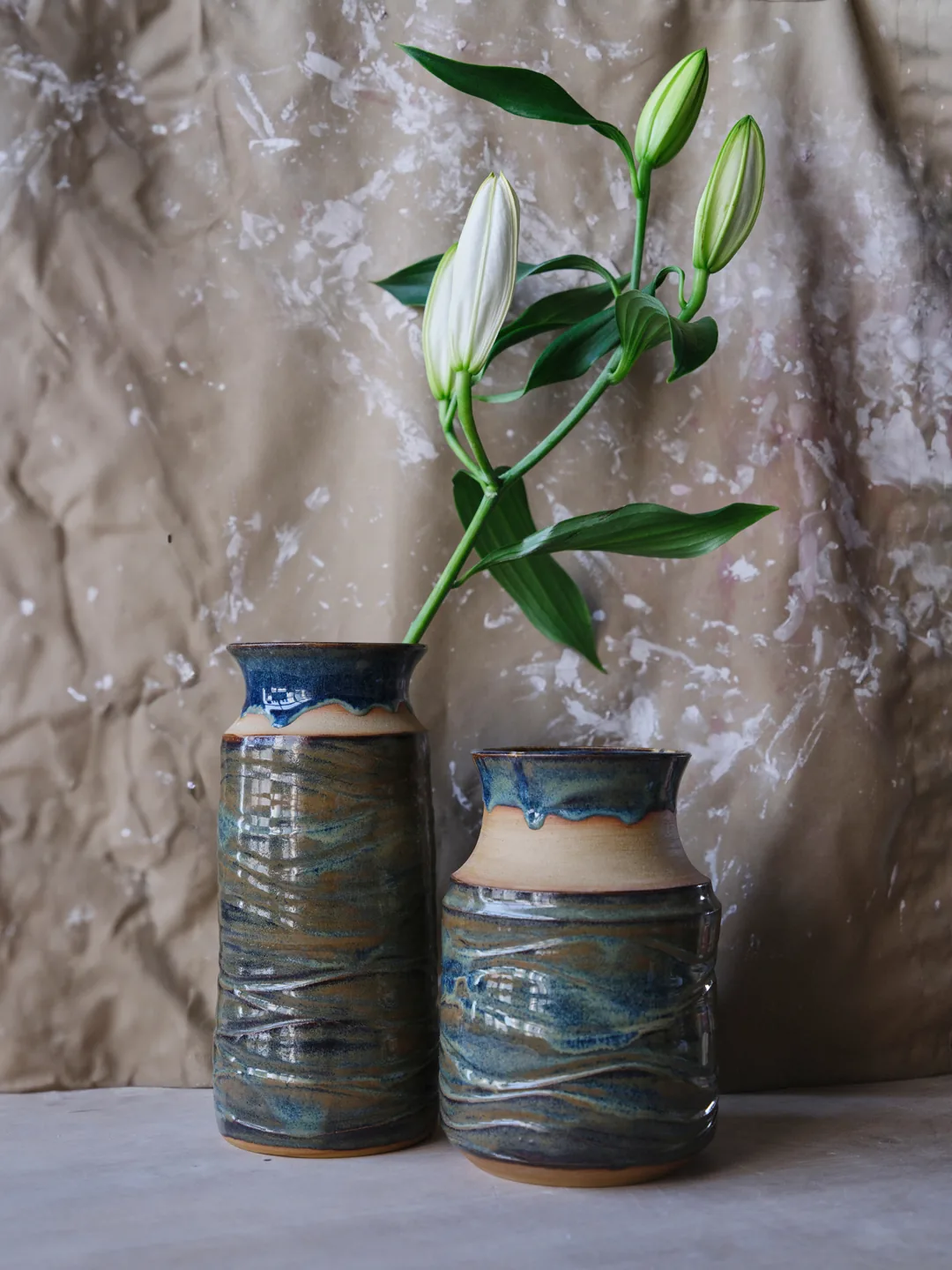 Two vases with a textured surface and a layered blue-green glaze, the taller one holding a stem of white lilies, against a brown paper backdrop.